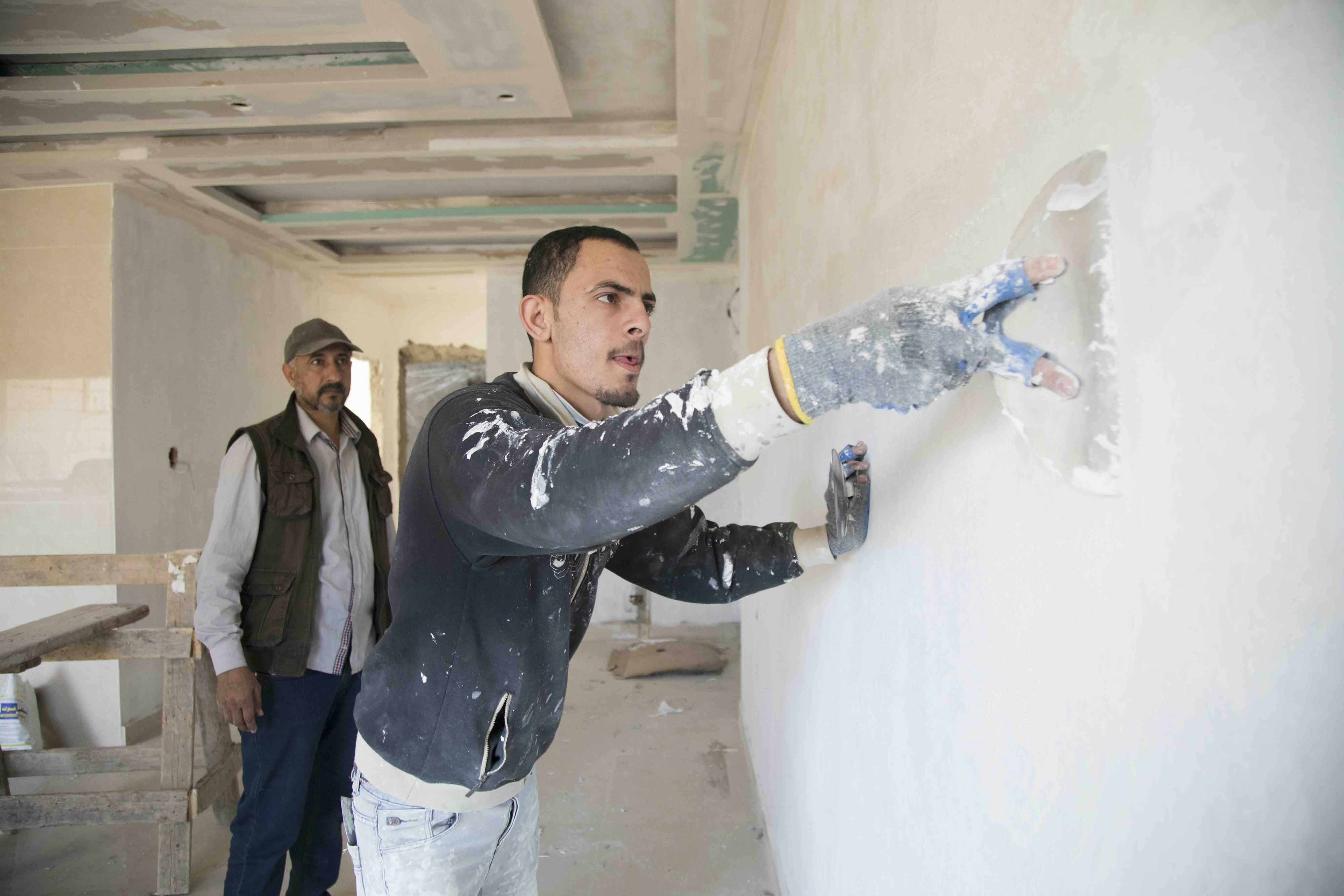 Image of a worker painting the wall in a room with another worker watching in the background