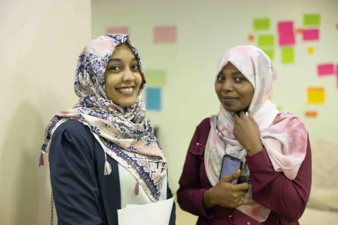 image of two female students in a conversation with post-it notes on a whiteboard in the background